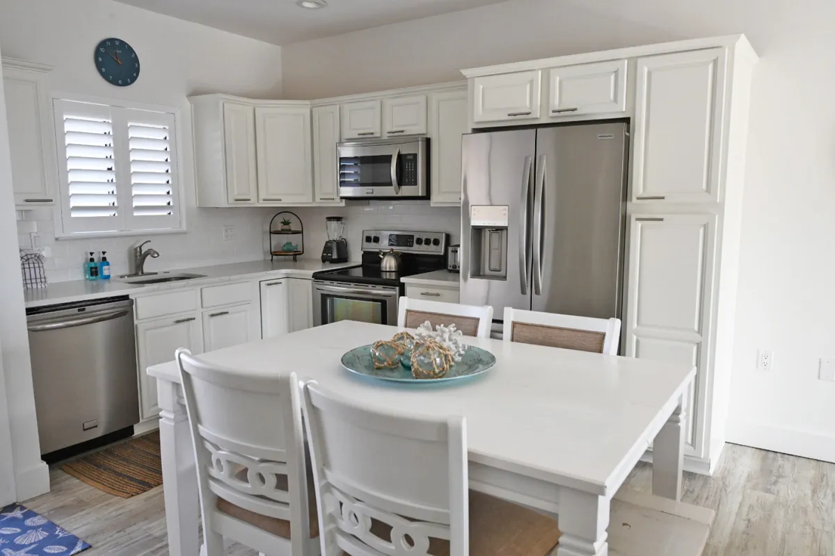A sun-drenched dining area featuring a modern dining table, crisp white cabinetry, and stainless steel kitchen appliances. A decorative bowl adds a touch of hominess to the welcoming, open-concept space. Natural light fills the room, enhancing its inviting atmosphere.