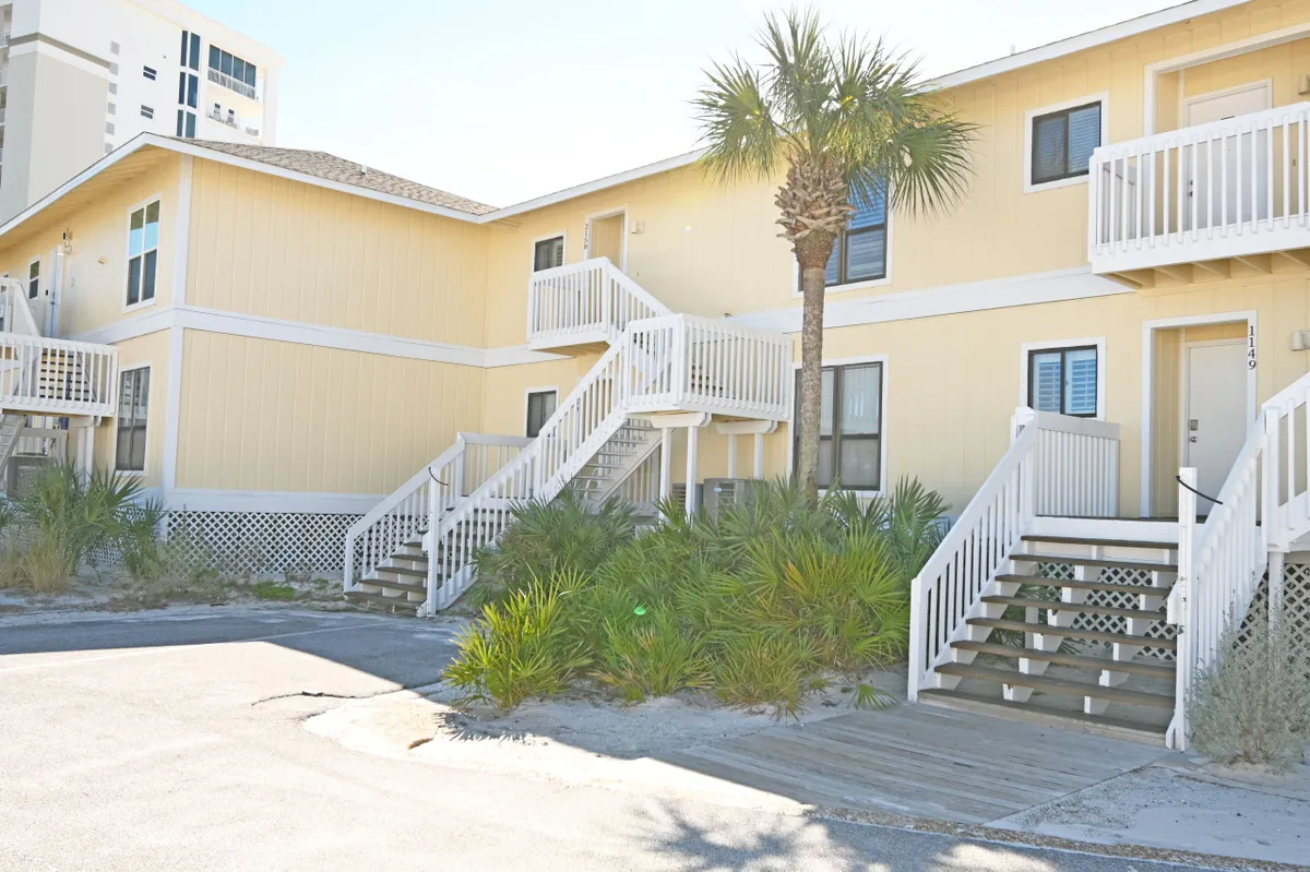 This exterior view shows the yellow building facade with stairs leading up to entrances and a palm tree in the foreground. The yard area at the base of the stairs offers a casual, friendly vibe well-suited to mingling with neighbors or letting pets explore under supervision.