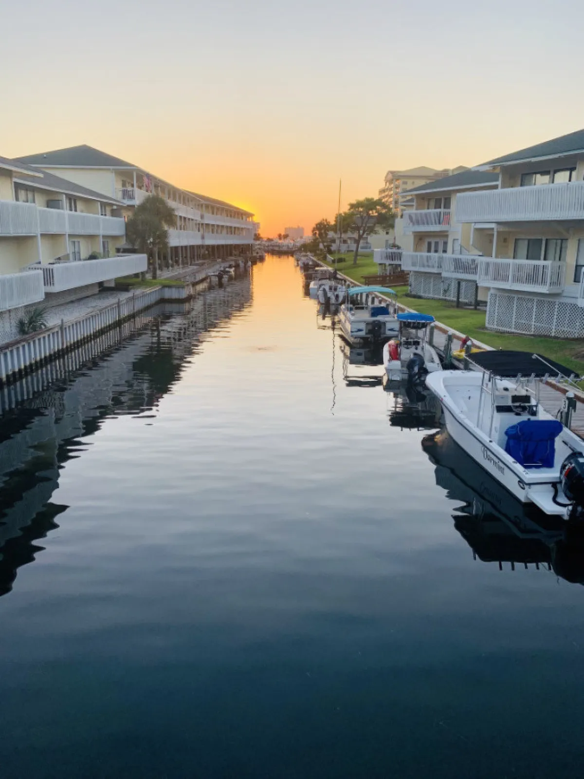 A serene waterway scene at sunset, with boats moored along the shore and buildings glowing in the golden hour. The tranquil waters and gentle sky evoke peaceful evening strolls, community events, and the charm of local festivals or boat parades.