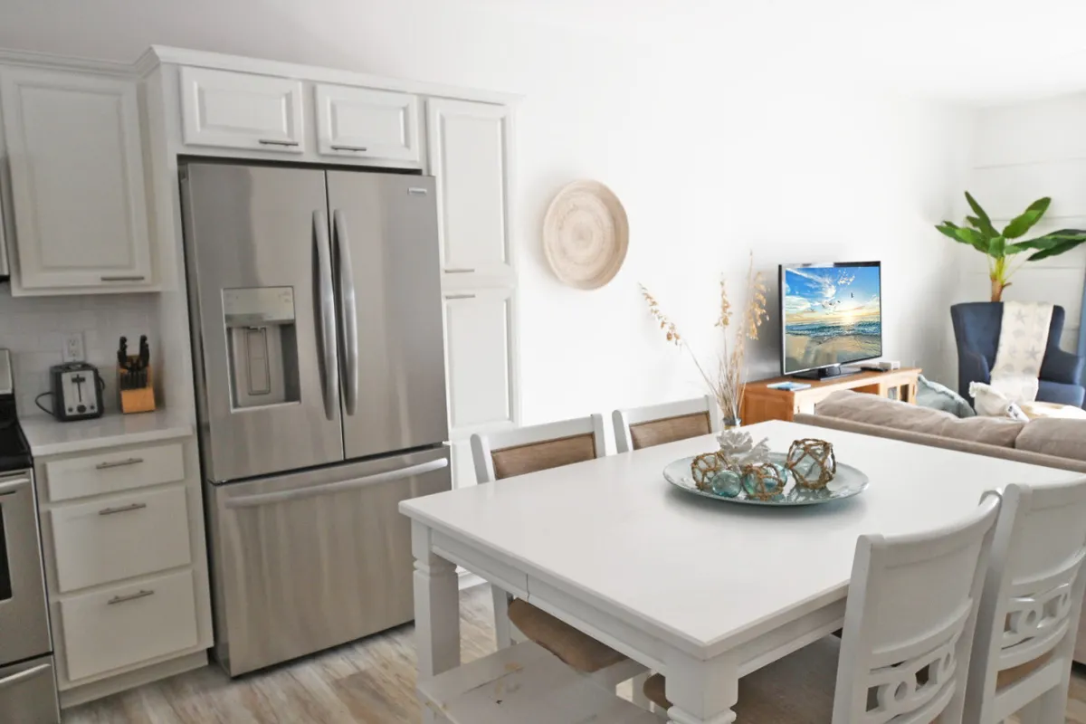 A view of the kitchen and dining area with a stainless steel refrigerator, a white dining table set for meals, and a television displaying a beach scene in the background. Lush indoor plants and a decorative bowl add to the inviting vibe, and the open layout hints at space for easy movement between the kitchen, dining area, and living space.