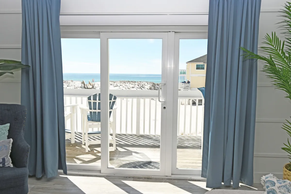The image showcases a sunlit interior living space leading to a spacious balcony through large sliding glass doors. Two inviting white Adirondack chairs sit on the balcony, overlooking a sweeping panoramic view of the turquoise Gulf of Mexico. The scene evokes a sense of tranquility and relaxation, with the gentle light and endless ocean horizon visible from both indoors and out.