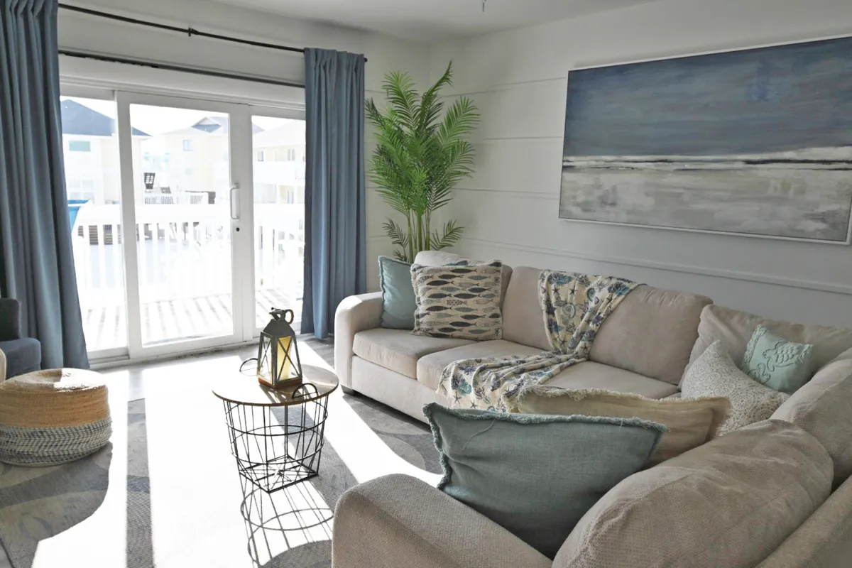 A cozy living area showcasing a large sectional couch adorned with beach-themed pillows, a wireframe coffee table with a lantern centerpiece, and expansive glass sliding doors that lead to a balcony. Natural light fills the space, and a large piece of ocean-inspired wall art emphasizes the coastal setting.