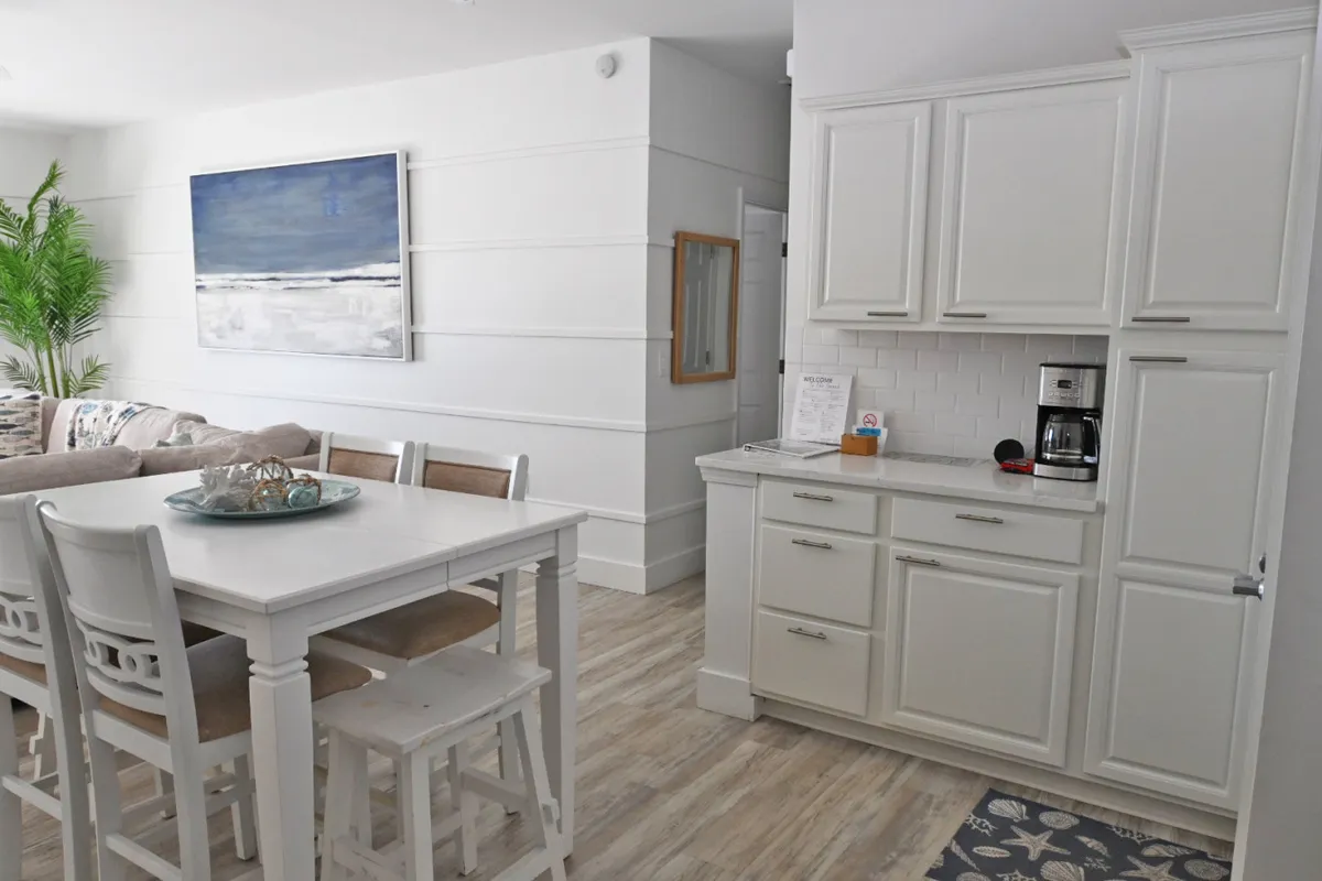 This inviting interior scene highlights a white dining table with chairs perfectly positioned near the kitchen's white cabinetry. A coffee maker is visible on the counter, signaling the dedicated coffee bar area. Large coastal artwork and a glimpse of the comfortable sectional couch complete the open-concept space, which is bathed in natural light, creating a fresh, coastal ambiance.