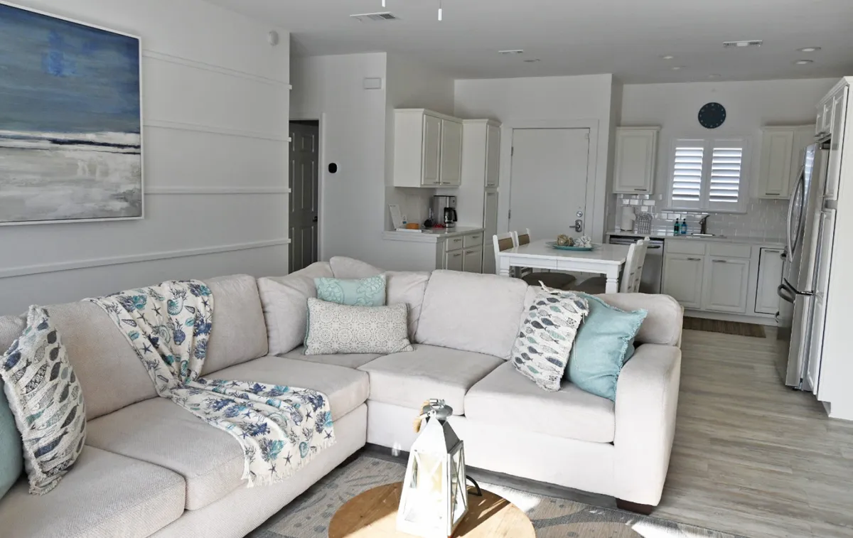 The photo captures a spacious, modern kitchen with stainless steel appliances and white cabinetry, seamlessly flowing into the living area. A large beige sectional couch with patterned pillows sits nearby, and the kitchen is clearly equipped for cooking and entertaining. Decorative coastal art adds to the beachy feel, while abundant natural light enhances the fresh, airy environment.