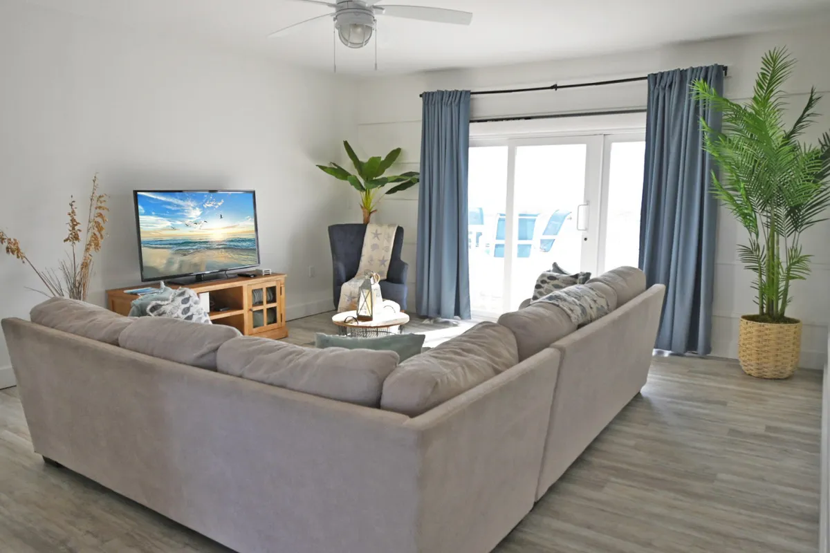 A bright, sunlit living room featuring a large beige sectional couch facing a smart flat-screen TV that displays a beach scene. Sliding glass doors with blue curtains open directly to a private balcony, and green potted plants add a touch of nature. The space exudes a calm, cozy, and refreshing coastal vibe.