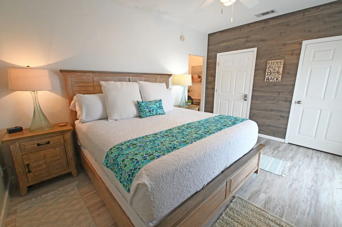 This image features a king-size bed dressed in crisp white linens with a teal accent runner, flanked by matching wooden nightstands and glass-base lamps. A wood accent wall and beach-themed sign add warmth and personality, while the overall ambiance is soothing and welcoming.