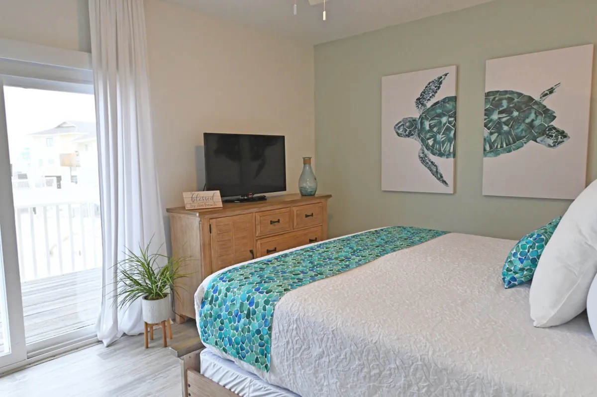 A peaceful guest bedroom with a bed featuring a turquoise and blue patterned runner and pillow, complemented by sea turtle wall art. The room includes a wooden dresser with a flat-screen TV and a sliding glass door leading to a private balcony, inviting natural light and fresh air.