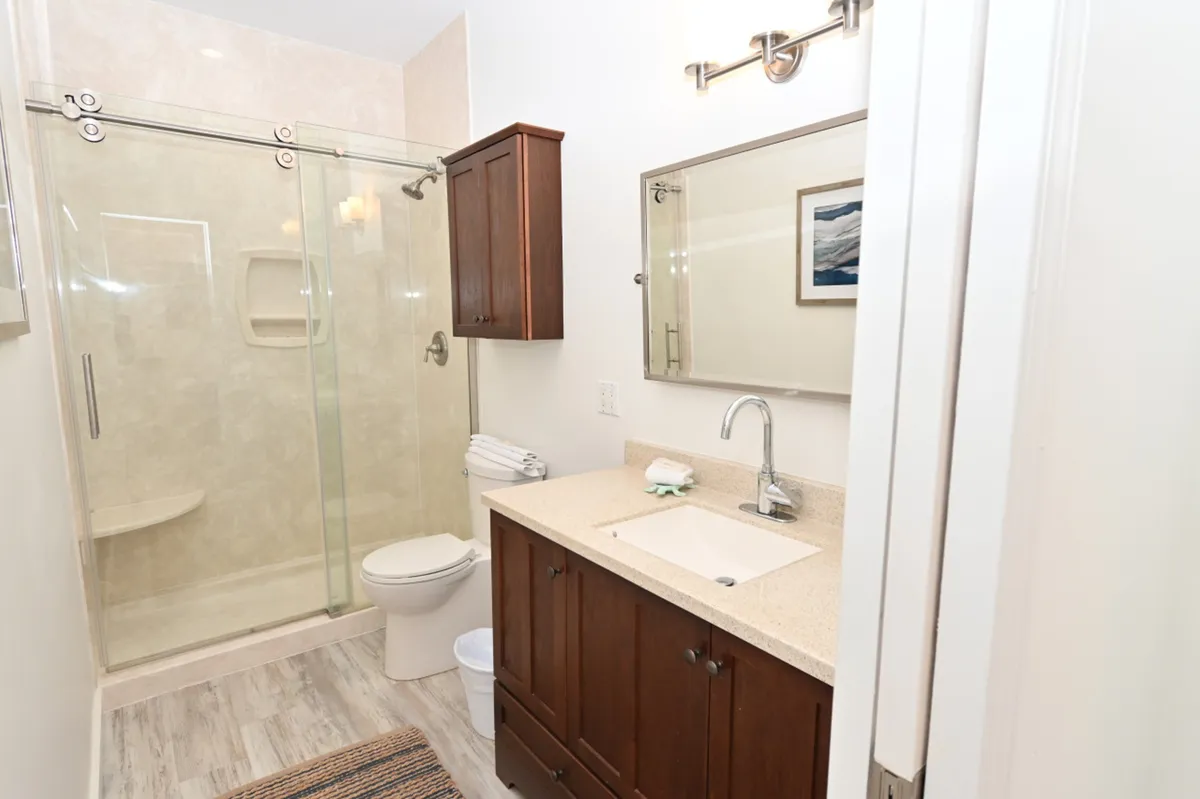 A modern bathroom with a glass-enclosed shower featuring a barn-style sliding door, a wooden vanity with a cream countertop and integrated sink, neatly folded plush towels, and a large mirror. The space is bright and exudes spa-like calm.