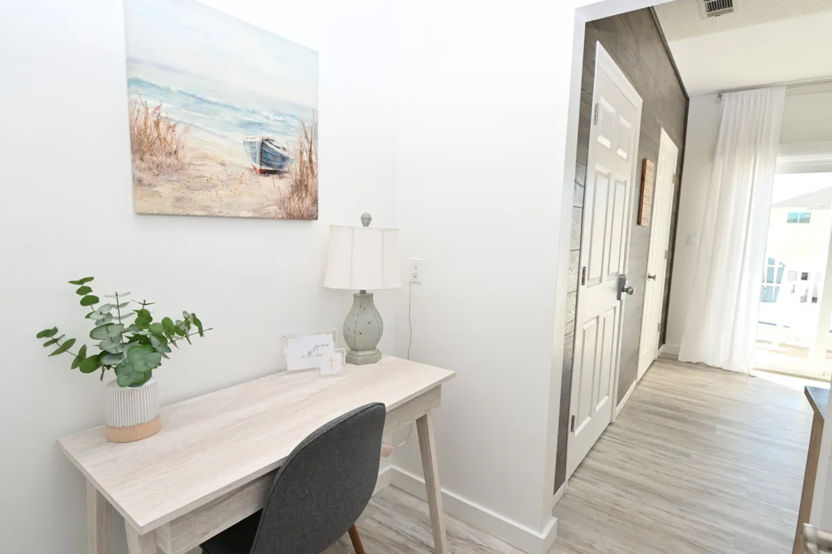 A simple, light wood desk with a lamp and plant is positioned near a bright hallway leading to a balcony. A gray upholstered chair and nautical-themed wall art add subtle coastal charm. Natural light pours into the workspace, creating an inviting, focused environment with easy access to the outdoors.
