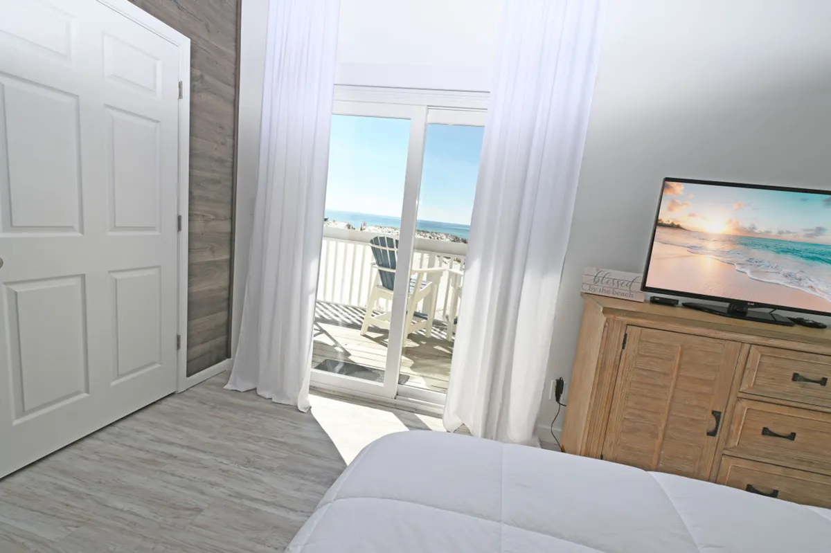 This image features a master suite with a sliding glass door that opens to a balcony overlooking the ocean. The room includes a smart TV, a rustic wooden dresser, white bed linens, and curtains that allow in abundant natural light. The inviting balcony, visible through the doors, hints at a serene place to take a break or get inspired by Gulf views.