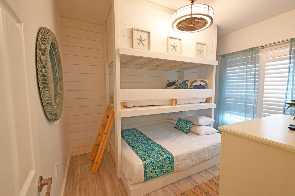 A cozy nook with a white bunk bed adorned in teal and blue patterned bedding, decorative starfish art above the bunks, and soft natural light filtering through blue sheer curtains and white shutters. The space feels playful and welcoming for children or groups.