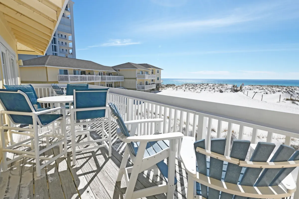 A private balcony with white wooden railing overlooks a pristine white sandy beach and the expansive blue ocean. Blue cushioned patio chairs and a round white table invite relaxation and morning coffee, while the horizon promises breathtaking sunrise or sunset moments. The scene is tranquil, airy, and utterly luxurious, placing the guest at the edge of the Gulf's beauty.