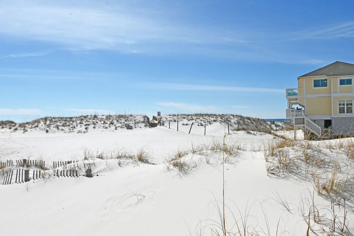 An exterior view of a tranquil white sand dune pathway leading to the beach, with a yellow beachfront house and balconies in the background. Sparse beach grasses and expansive coastal views evoke relaxation and a sense of escape.