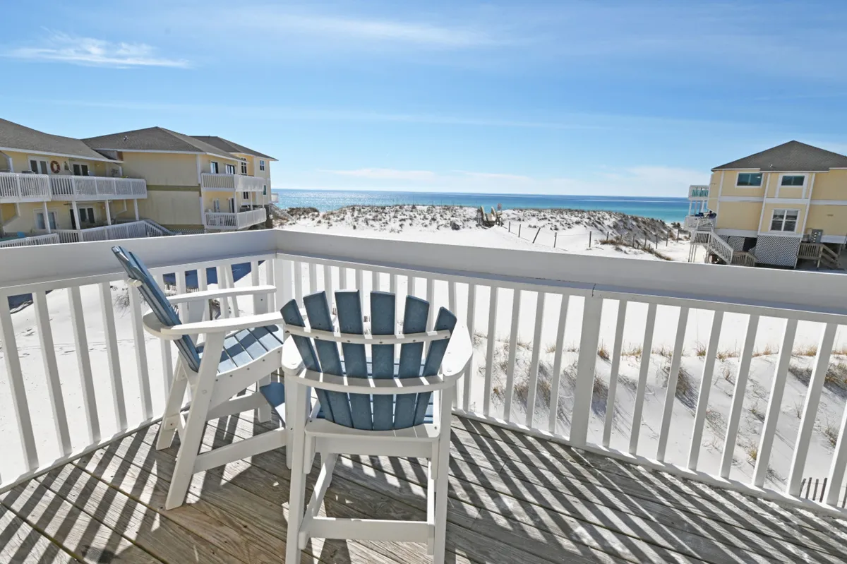 Two blue and white Adirondack chairs on a private balcony overlook pristine white sand dunes and the blue-green Gulf of Mexico. The white balcony railing casts soft shadows, and the scene is imbued with the tranquility of early morning or late afternoon light.