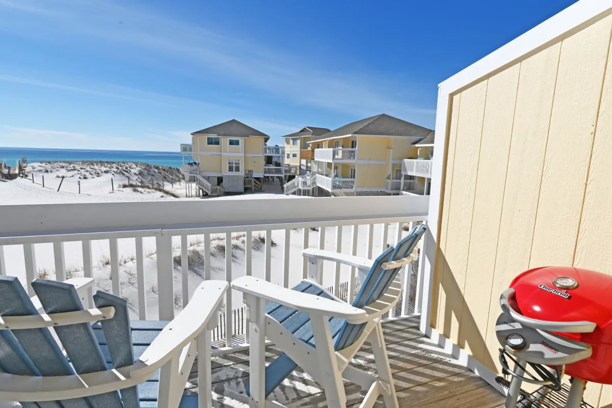 This exterior shot features two Adirondack-style chairs and a red charcoal grill on the private balcony, which overlooks sandy dunes and the distant beach. The white railing and yellow beach houses in the background set a tranquil, vacation-ready scene, inviting guests to enjoy outdoor dining or leisurely moments with beautiful coastal views.