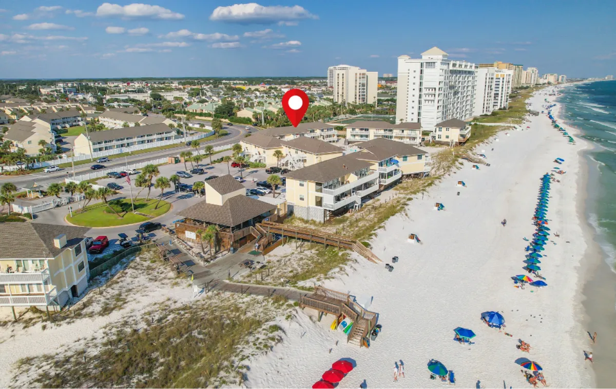 A vibrant beachfront scene featuring Sandpiper Cove’s condominium buildings directly facing the pristine white sand and emerald waters of the Gulf of Mexico. Colorful umbrellas dot the beach, and wooden boardwalks provide easy access to the shoreline. The lively atmosphere suggests families sunbathing, swimming, and engaging in outdoor games like pickleball and tennis, with the community pool and courts nearby.
