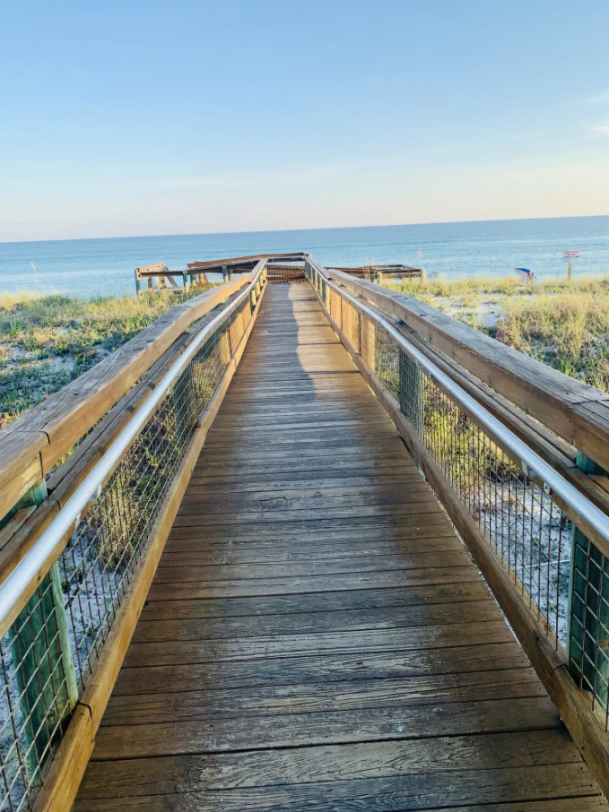 A serene wooden boardwalk leads through grassy dunes to an expansive view of the Gulf of Mexico. The scene is calm and rejuvenating, with the natural beauty of the coast on full display. This photo evokes off-season tranquility, inviting guests to imagine peaceful walks and sunset viewing along the shoreline.