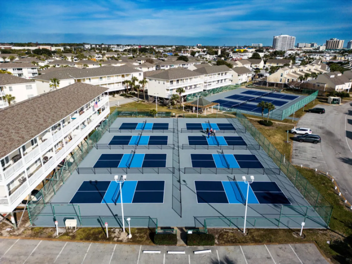Multiple blue and gray pickleball courts are set beside residential buildings and parking areas, with bright light poles indicating evening play possibilities. The courts are surrounded by open spaces for walking or jogging, creating a lively, active atmosphere.