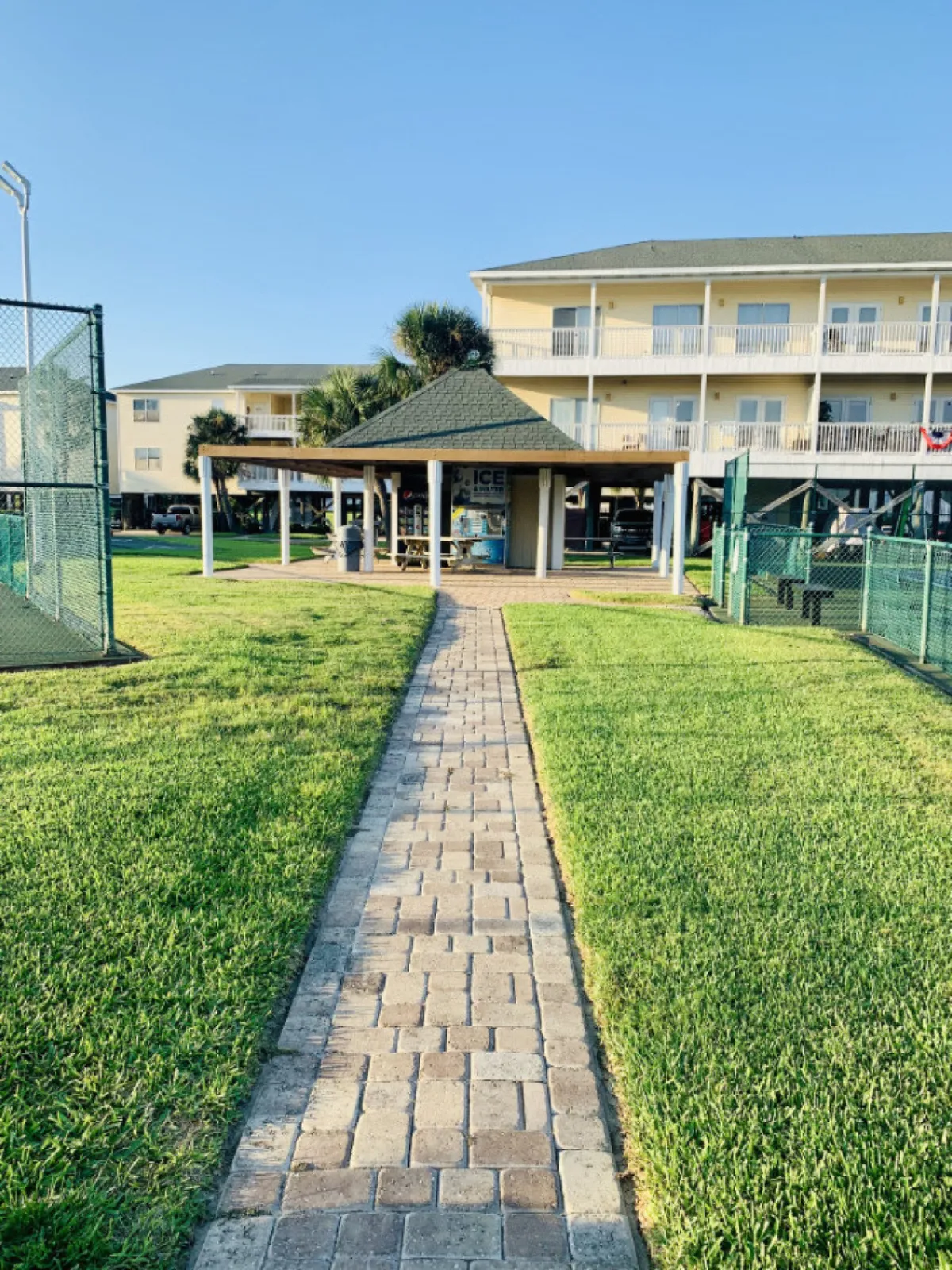 This image features a brick pathway leading to a covered pavilion with picnic tables, adjacent to tennis courts enclosed by green fencing. The scene is lively and communal, with a three-story yellow condominium building and balconies visible in the background, suggesting a vibrant, active environment within the Sandpiper Cove community.