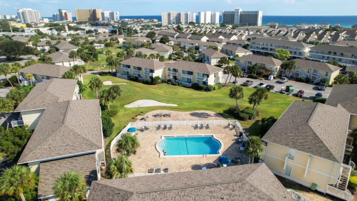 A cheerful exterior view of Sandpiper Cove’s central heated swimming pool, surrounded by lounge chairs and palm trees. Rows of townhouse-style vacation condos with balconies frame the pool, while the Gulf of Mexico glimmers in the background. The scene evokes a resort-like setting perfect for both family relaxation and play, with easy access from units to poolside fun.