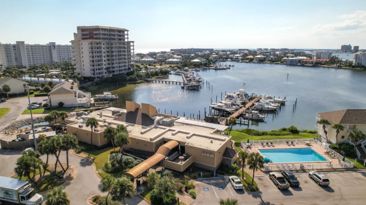 This image captures a marina with docked boats, a community swimming pool, nearby residential buildings, and a parking lot. The calm water and inviting pool area convey a relaxed yet active waterfront lifestyle.
