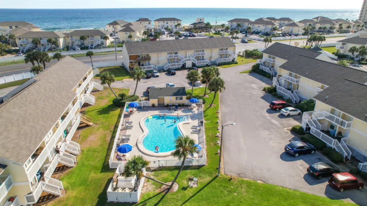 A central swimming pool with lounge chairs and umbrellas is surrounded by yellow two-story condominium buildings with private balconies. The background reveals a glimpse of the Gulf of Mexico, tying together the communal leisure spaces with the coastal setting. The scene is lively yet still relaxed, suggesting both social and solitary moments of comfort and enjoyment.