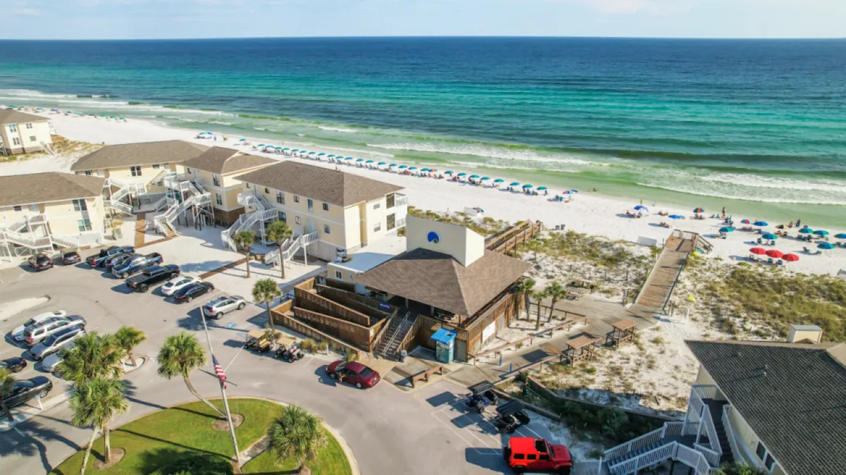 A pristine white sandy beach with colorful umbrellas stretches alongside turquoise Gulf waters. Beachfront buildings with private balconies overlook the shore, while a boardwalk provides gated direct access to the sand and surf. The scene is lively, with guests enjoying sunbathing, swimming, and the benefits of free beach service.