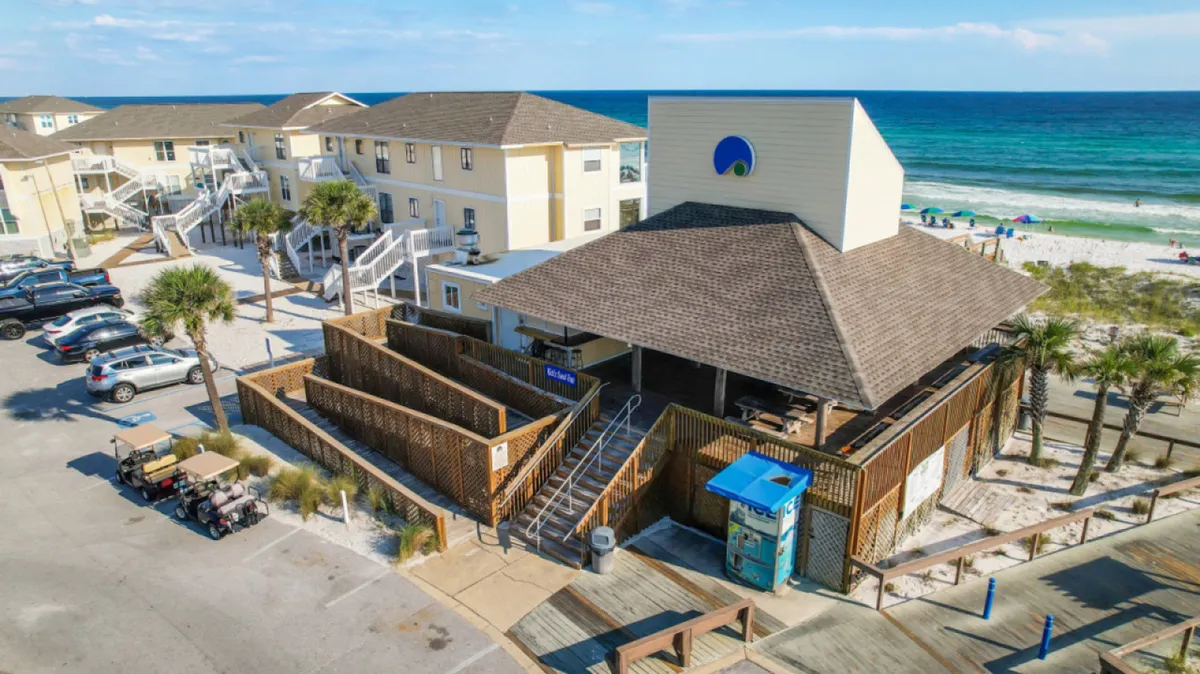 This image captures a vibrant beachfront scene, featuring a building with a large covered patio overlooking a pristine white sand beach and turquoise waters. The area is lively, with visible beach amenities such as lounge chairs and umbrellas, hinting at the complimentary beach service mentioned in the section. The overall mood is inviting and relaxing, perfectly embodying the quintessential Destin beach experience.