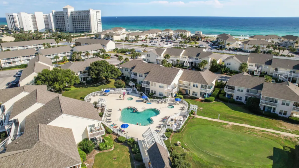 A central, kidney-shaped swimming pool with sapphire water is surrounded by lounge chairs and umbrellas. Clusters of two-story residential buildings with beige siding and brown roofs encircle the pool, with the Gulf of Mexico visible in the background. Well-maintained lawns and walking paths invite guests to enjoy the resort’s outdoor amenities.
