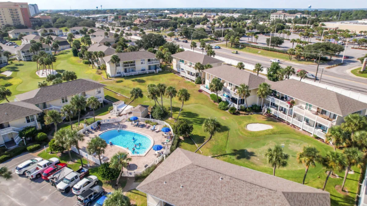 The community swimming pool area is surrounded by lounge chairs, umbrellas, lush green lawns, and palm trees. Low-rise condominium buildings with balconies provide the backdrop, and amenities like a putting green are visible. The overall atmosphere is casual and leisurely, emphasizing the year-round availability of resort amenities.