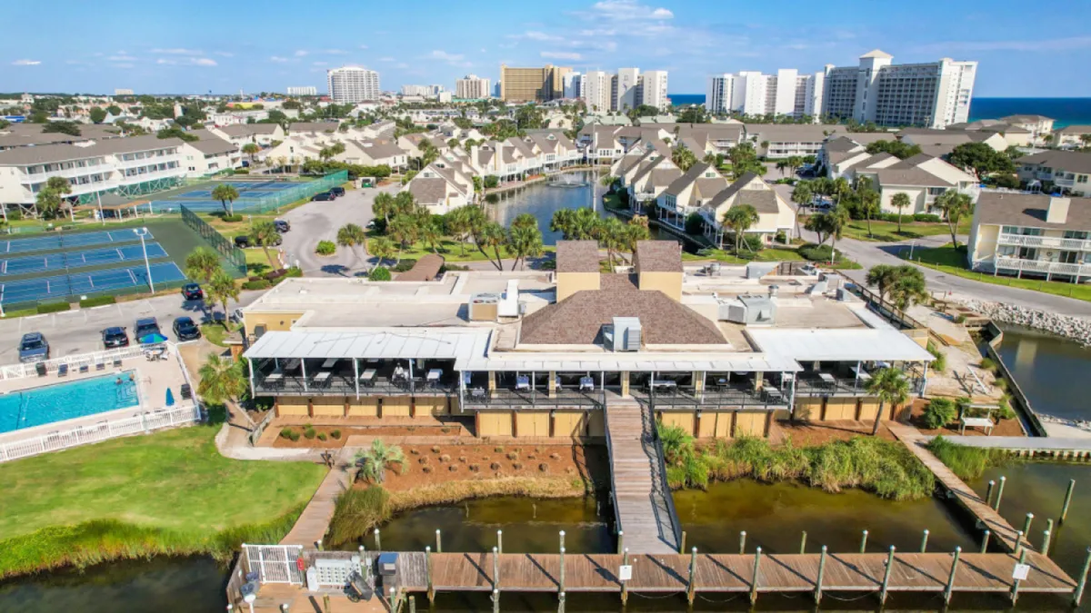 This exterior image showcases the community clubhouse with covered outdoor seating, adjacent tennis courts, a sparkling swimming pool, and marina docks with boat slips. The setting is sunny and vibrant, ideal for a variety of water and land-based activities.